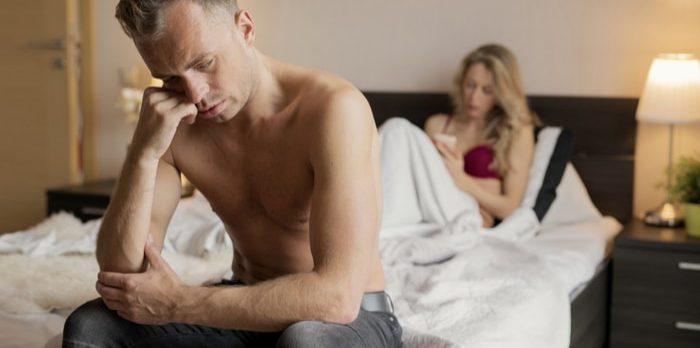 sexually incompatible - A lady under the sheets stting on the bed while using her cellphone. A man sitting at the edge of the bed looking worried.